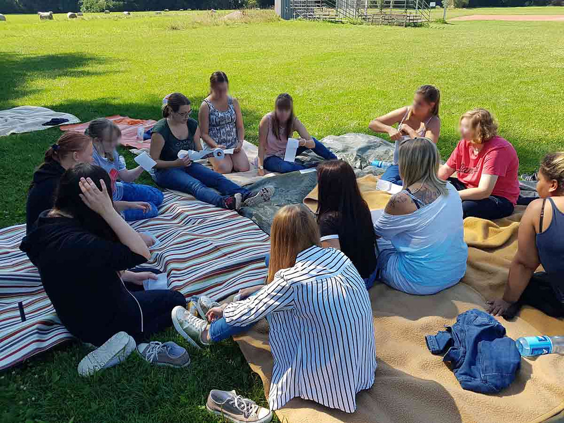 Wo Lernen lebendig wird Eine Gruppe junger Erwachsener sitzt im Kreis auf einer Wiese während eines Workshops zur Teamentwicklung und tauscht sich über ihre Aufgaben aus.