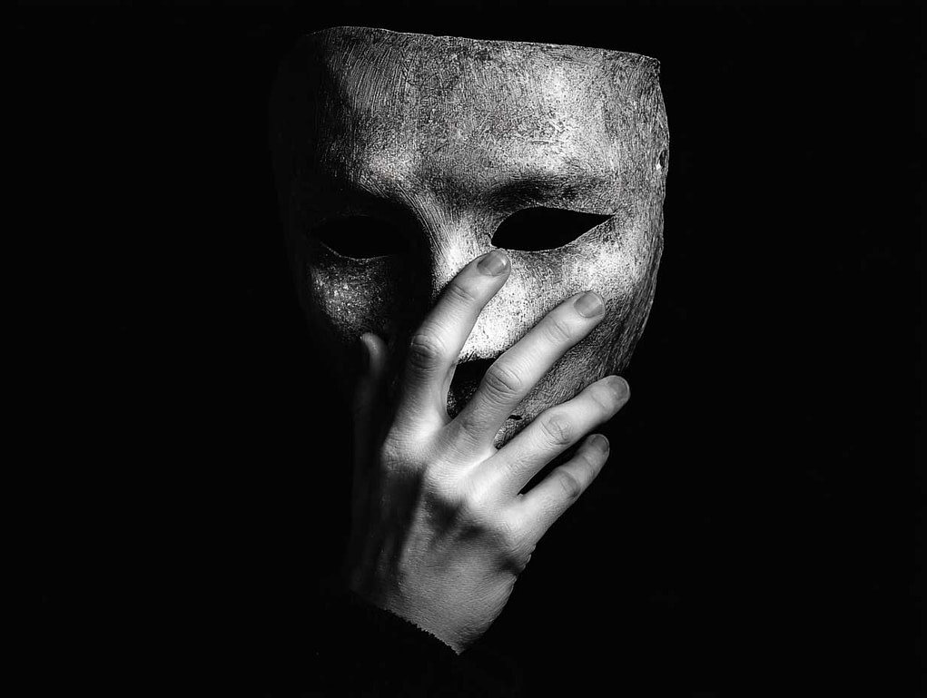 Kontrastreiche Schwarz-Weiß-Fotografie einer Maske und einer Hand – versinnbildlicht die kognitive Steuer (Covering) und die Last der Unfehlbarkeit.