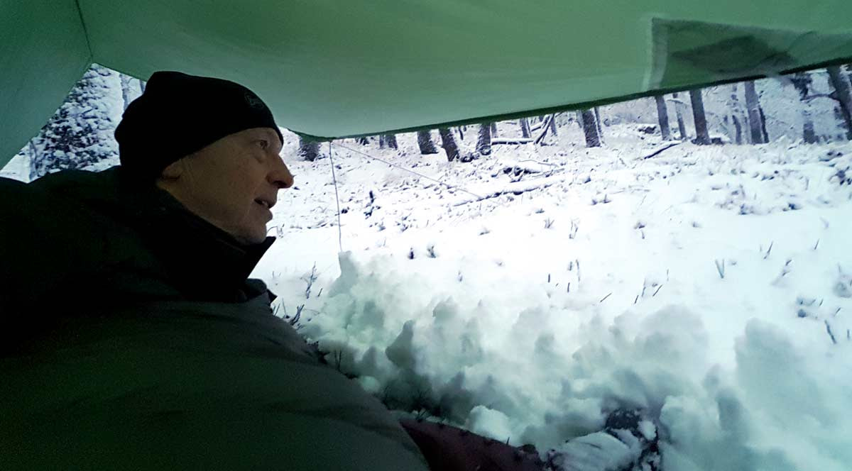raunaechte-hochsensibel-winterbiwak-schnee Ein Mann (Uwe Kreß) liegt im Winterwald in einem Schlafsack direkt auf dem Boden und ist von einer dicken Schicht Neuschnee bedeckt. Das Bild symbolisiert Rückzug, Kälteanpassung und die psychologische Erfahrung der Raunächte für Hochsensible.