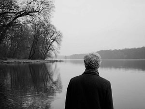 Ein reifer Mann blickt ruhig über einen weiten See – Symbol für die Stille nach dem Lärm und das Ablegen der Jupiter-Maske.