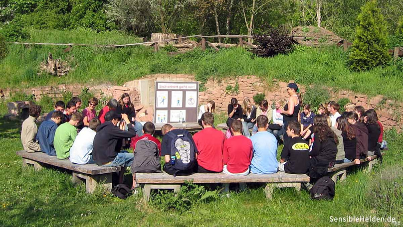 Schueler-Reflexion-Hochseil Reflexionsrunde im Hochseilgarten Worms – Schülergruppe im Sitzkreis mit Trainerin, im Hintergrund die Tafel Sicherheit geht vor