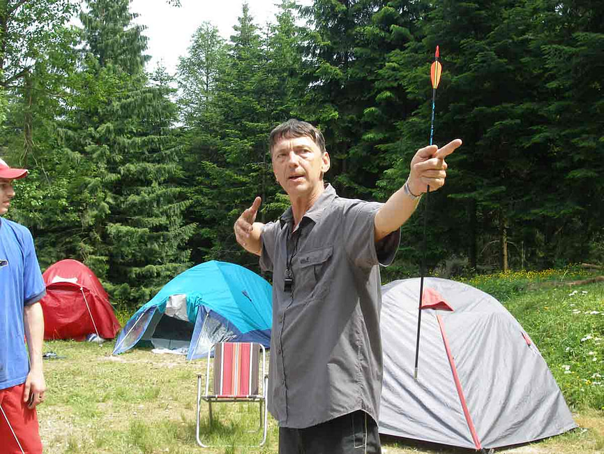 Trainer Götz Uwe Kreß steht in einem Zeltlager im Wald und erklärt einer Gruppe mit einem Pfeil in der Hand eine Übung zur Persönlichkeitsentwicklung.